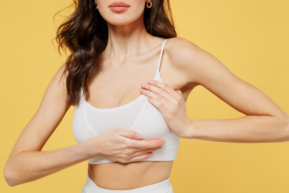 A woman in a white sports bra against a yellow background, showcasing the improved shape, firmness, and elevated nipple position of a natural-looking breast lift.